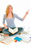 Smiling teen girl sitting on floor among schoolbooks and pointing in corner
