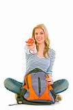Cheerful teenager sitting on floor with schoolbag and giving apple
