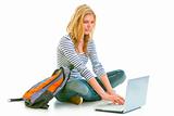 Pensive teenager sitting on floor with backpack and using laptop
