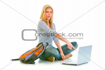 Surprised teenager sitting on floor with backpack and pointing in laptop
