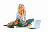 Tired teen girl sitting on floor with backpack and looking on laptop
