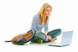 Interested pretty girl sitting on floor with backpack and looking on laptop
