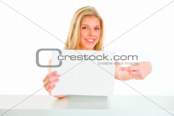 Smiling girl sitting at table and pointing on blank paper

