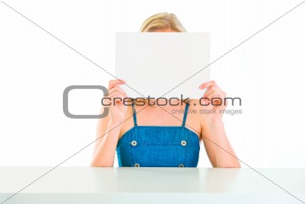 Girl sitting at table and holding blank paper in front of face
