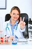 Smiling doctor woman sitting at office table and showing victory gesture
