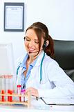 Smiling doctor woman with headset sitting at office table and talking with someone
