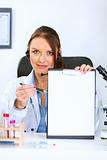 Doctor woman sitting at office table showing blank clipboard and pen for signing
