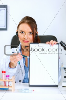 Doctor woman sitting at office table showing blank clipboard and pen for signing
