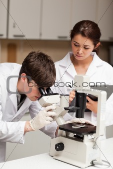 Portrait of a scientist looking in a microscope while his colleague is taking notes