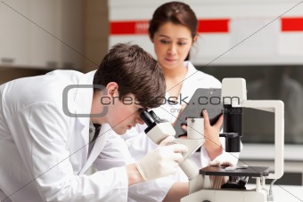 Scientist looking in a microscope while his colleague is writing