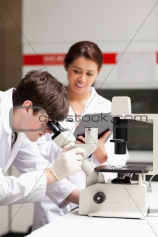 Portrait of a science student looking in a microscope while his classmate is writing