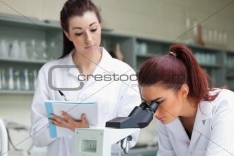 Student looking into a microscope while her classmate is taking notes