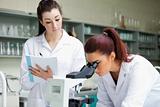 Scientist looking into a microscope while her colleague is taking notes