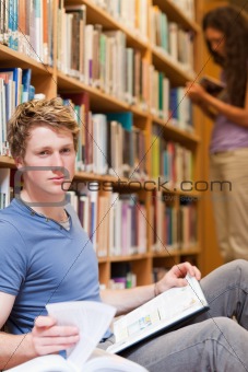 Portrait of a handsome student posing with a book while his classmate is reading