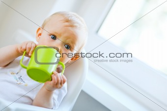 Pretty baby girl sitting in chair and drinking from baby cup 

