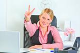 Smiling business woman sitting at office desk and  showing victory gesture
