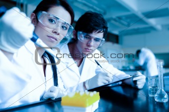 Science student dropping liquid in test tubes while her partner is taking notes