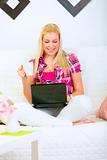 Happy modern housewife sitting on sofa with laptop and cup of coffee
