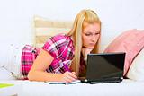 Concentrated young woman lying on couch with laptop and writing in notepad
