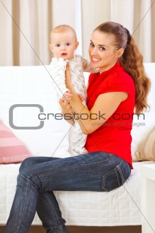 Happy mother sitting on divan and holding adorable baby in hands
