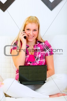 Happy young housewife sitting on divan with laptop and talking phone
