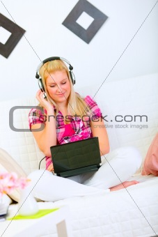 Happy young woman sitting on sofa with laptop and listening music in headphones
