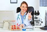 Smiling female medical doctor sitting at office table and showing victory gesture
