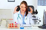 Stressed female medical doctor sitting at office table and holding hands near head
