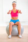 Happy young female sitting on fitness ball and showing thumbs up
