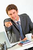 Smiling modern businessman sitting at office desk and pointing on you
