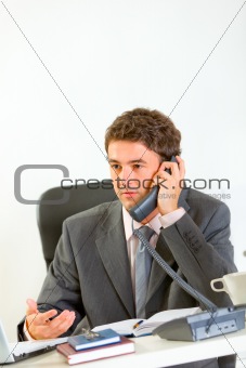 Serious modern businessman sitting at office desk and speaking phone
