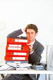 Confused businessman sitting at office desk with pile of folders
