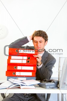 Confused businessman sitting at office desk with pile of folders
