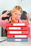 Stressed modern female manager with pile of folders at office desk
