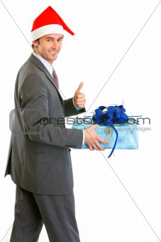 Happy modern businessman in Santa Hat holding present and showing thumbs up
