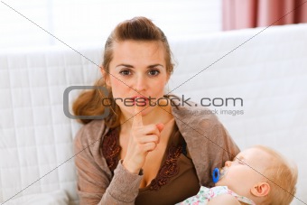 Young mama holding sleeping baby and showing shh gesture
