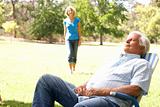 Senior Man Relaxing In Park With Wife In Background