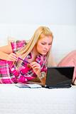Pretty young woman laying on couch with notepad and pen and looking on laptop

