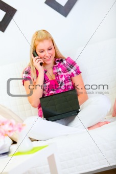 Smiling young woman sitting on couch with laptop and speaking mobile phone
