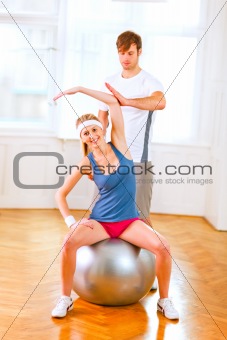 Smiling girl making exercises on fitness ball assisted by her personal trainer 
