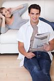 Man leaning against the sofa with newspaper while his girlfriend listens to music