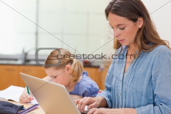 Little girl doing her homework while her mother is using laptop