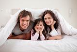 Smiling parents lying under a duvet with their daughter