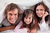 Happy parents lying under a duvet with their daughter