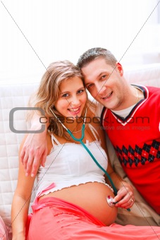 Smiling pregnant woman sitting on sofa with young husband and listening her tummy using stethoscope
