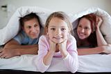 Cheerful girl under bed cover with parents