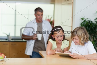 Children using tablet in the kitchen with father behind them
