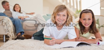 Siblings doing their homework on the floor with parents behind them