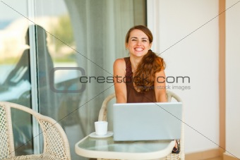 Happy young woman sitting on terrace with cup of coffee and laptop
