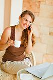 Smiling woman sitting on terrace with laptop and cup of tea and speaking mobile 
